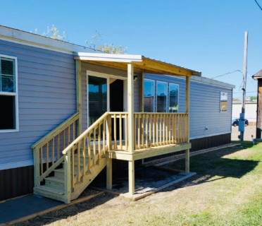 Small beige house with a wooden porch, steps, and railing on a sunny grassy lot.