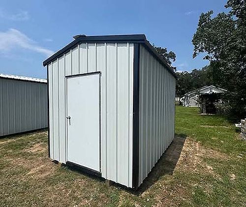 Small gray storage shed with white door on grass, outdoors under a blue sky.