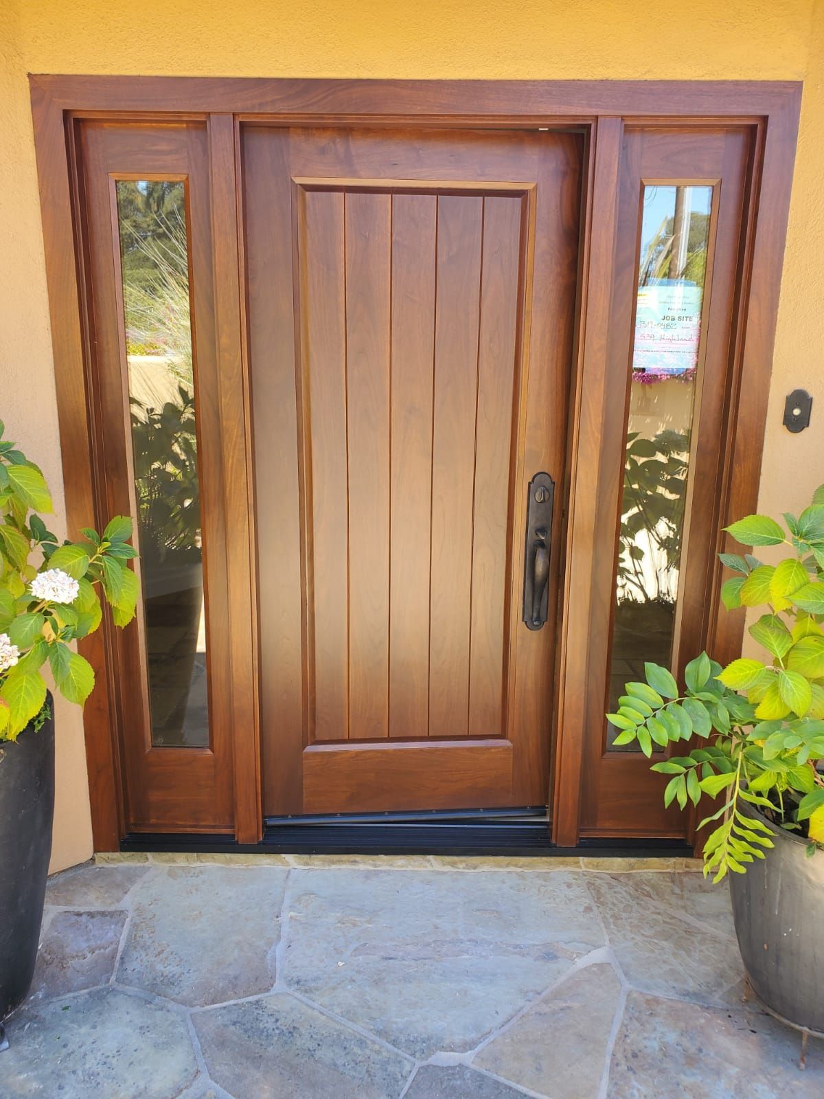 Wooden front door with glass side panels, black handles, and potted plants beside stone entryway.