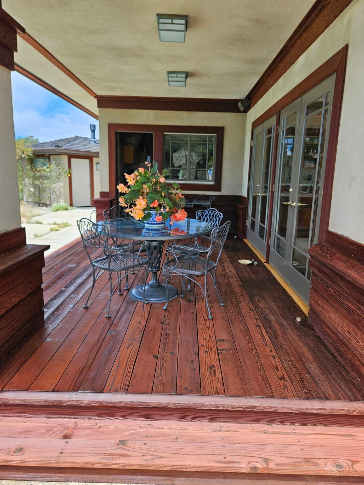 Wooden porch with a metal table and chairs, flower arrangement, and glass doors along the side.
