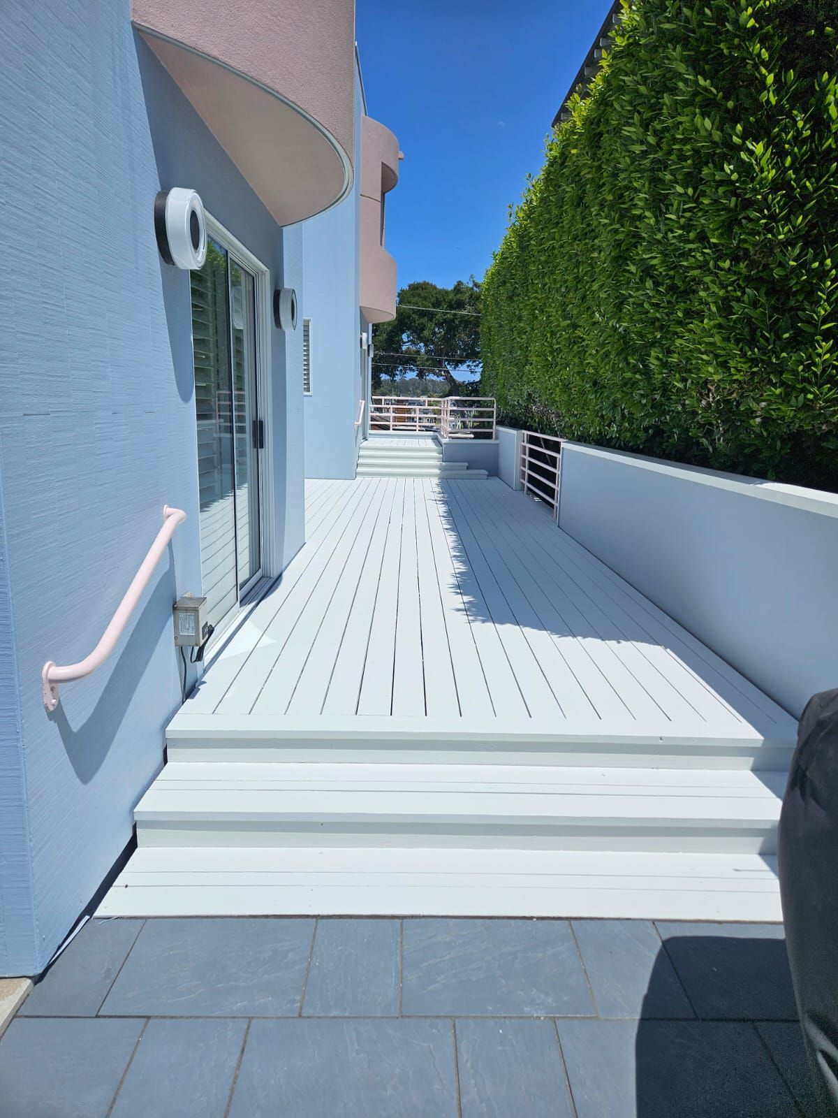 White wooden deck walkway beside a light blue house, bordered by a tall green hedge under bright sunlight.