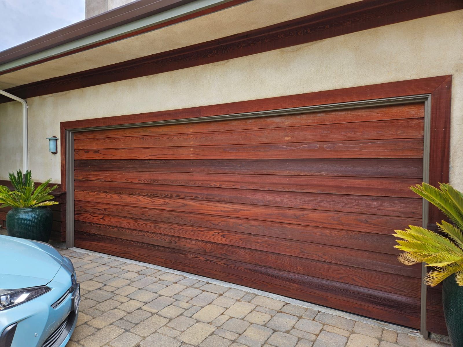 Modern reddish-brown garage door beside a light blue car in a paved driveway