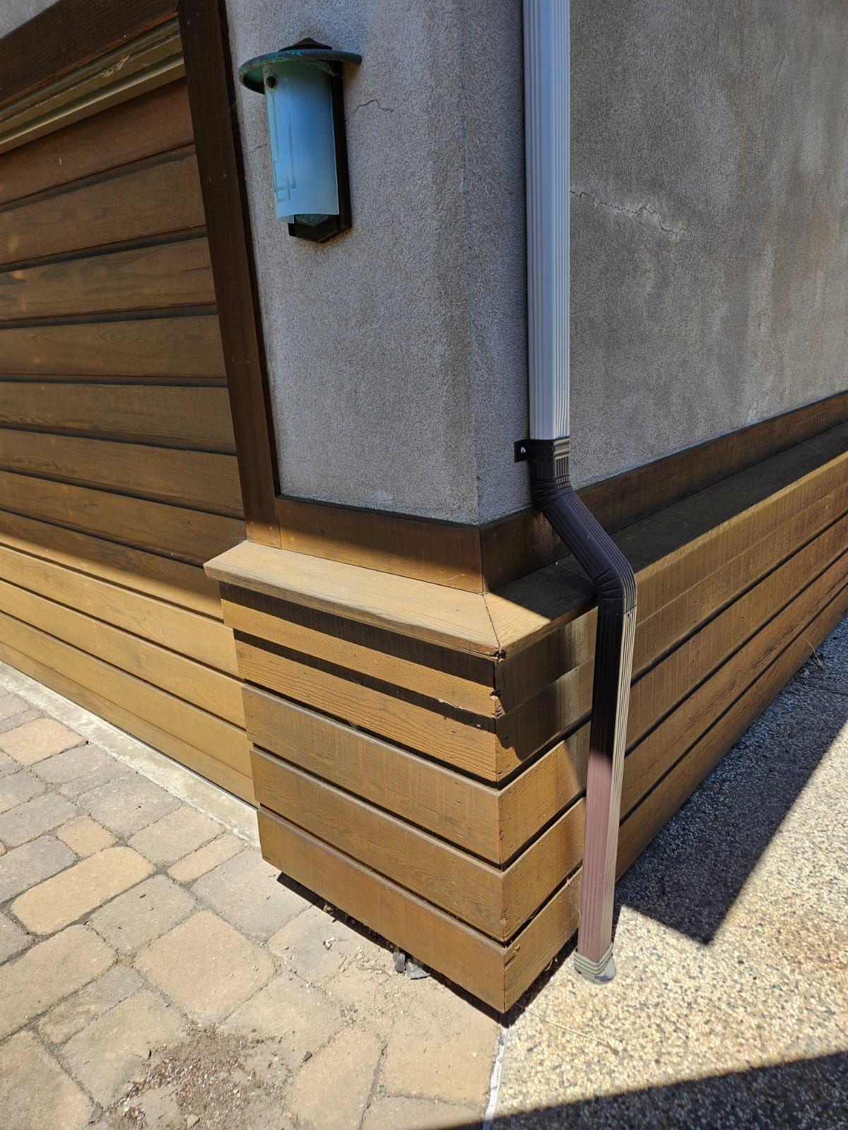 Building corner with wood paneling, gray stucco wall, downspout, and wall-mounted light.