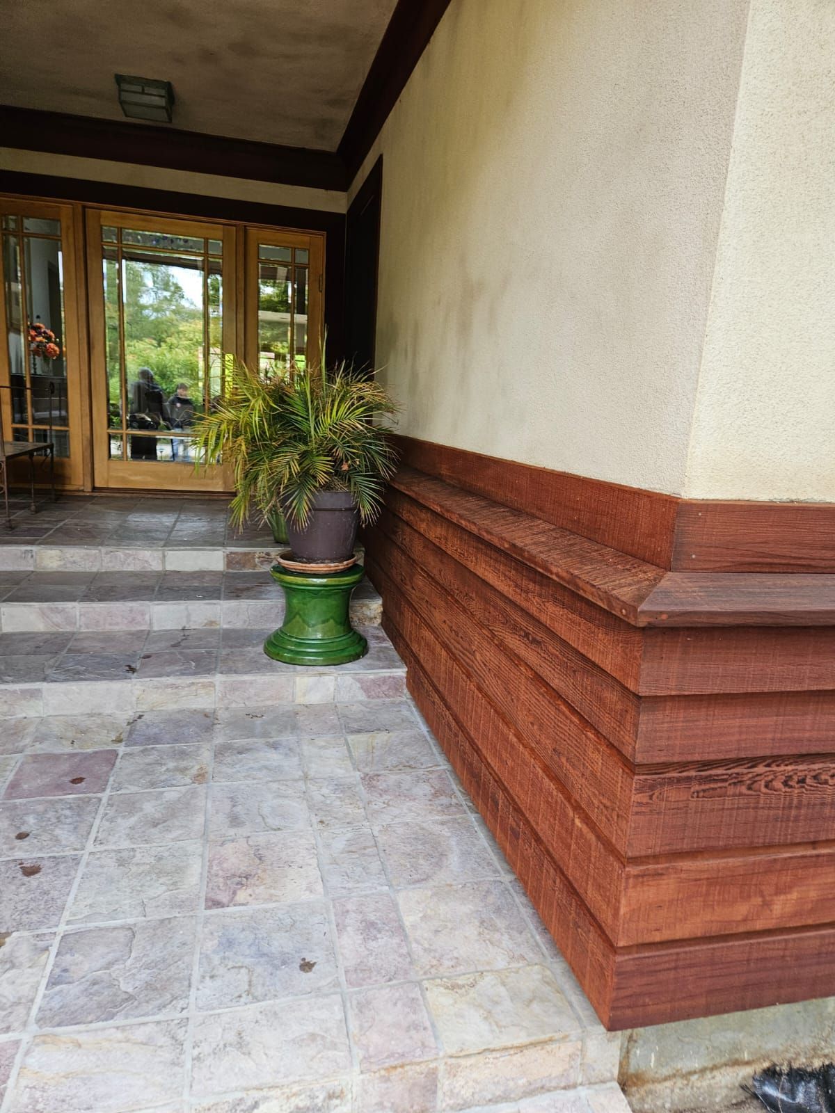 Covered porch with tiled floor, wooden bench-style wall trim, potted plant, and glass front doors.