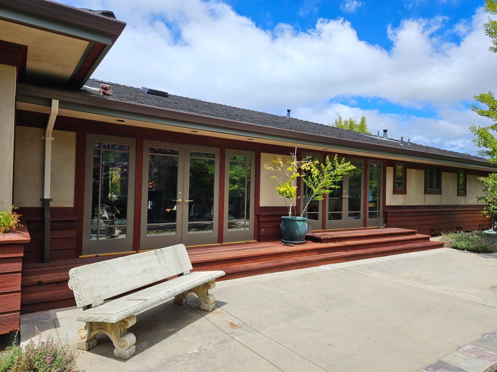 Modern house patio with wooden deck, glass doors, and a concrete bench under a blue sky