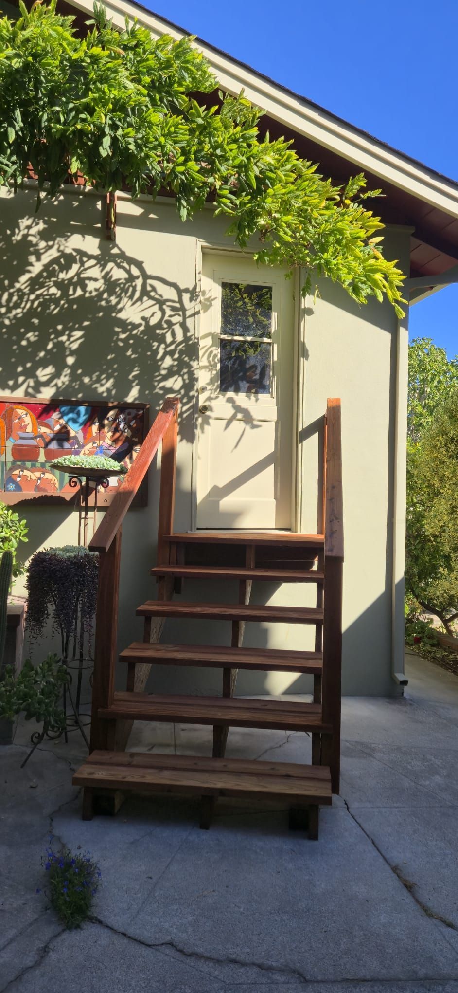 Wooden outdoor staircase beside a beige house, with leafy vines overhead and a cracked driveway below