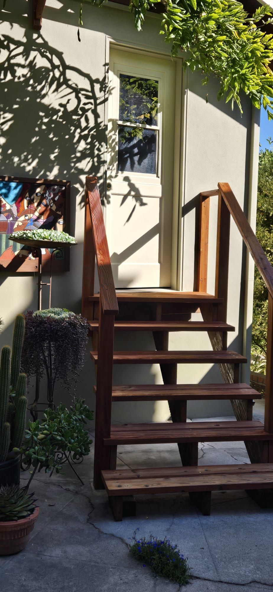 Wooden outdoor stairs with a handrail leading to a small cream-colored house porch in sunlight.