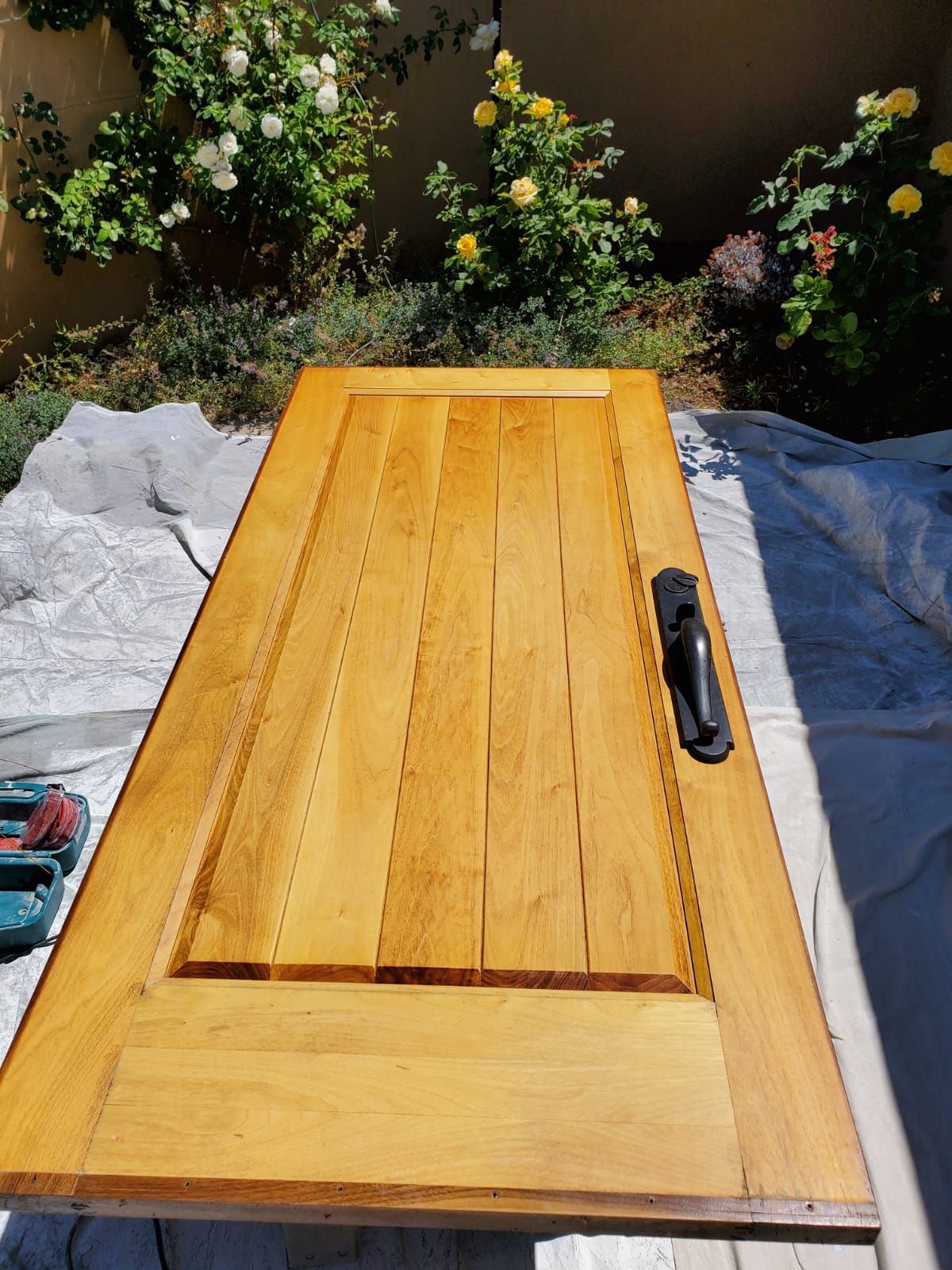 Wooden door panel laid flat outdoors on tarps beside flowers, with a tool resting on it.