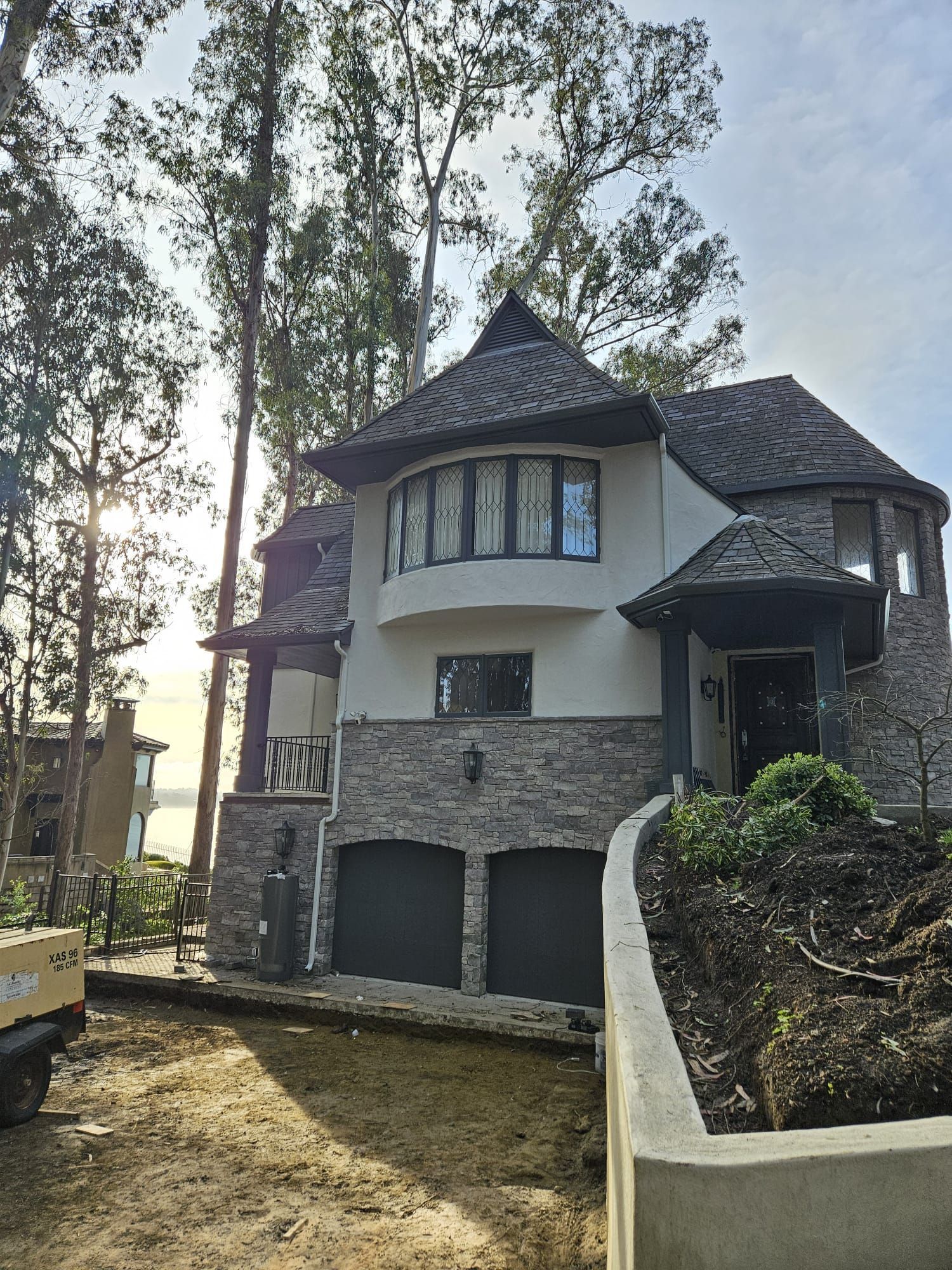 Stone-and-stucco house with dark roof and bay window, viewed from a dirt driveway beside tall trees.