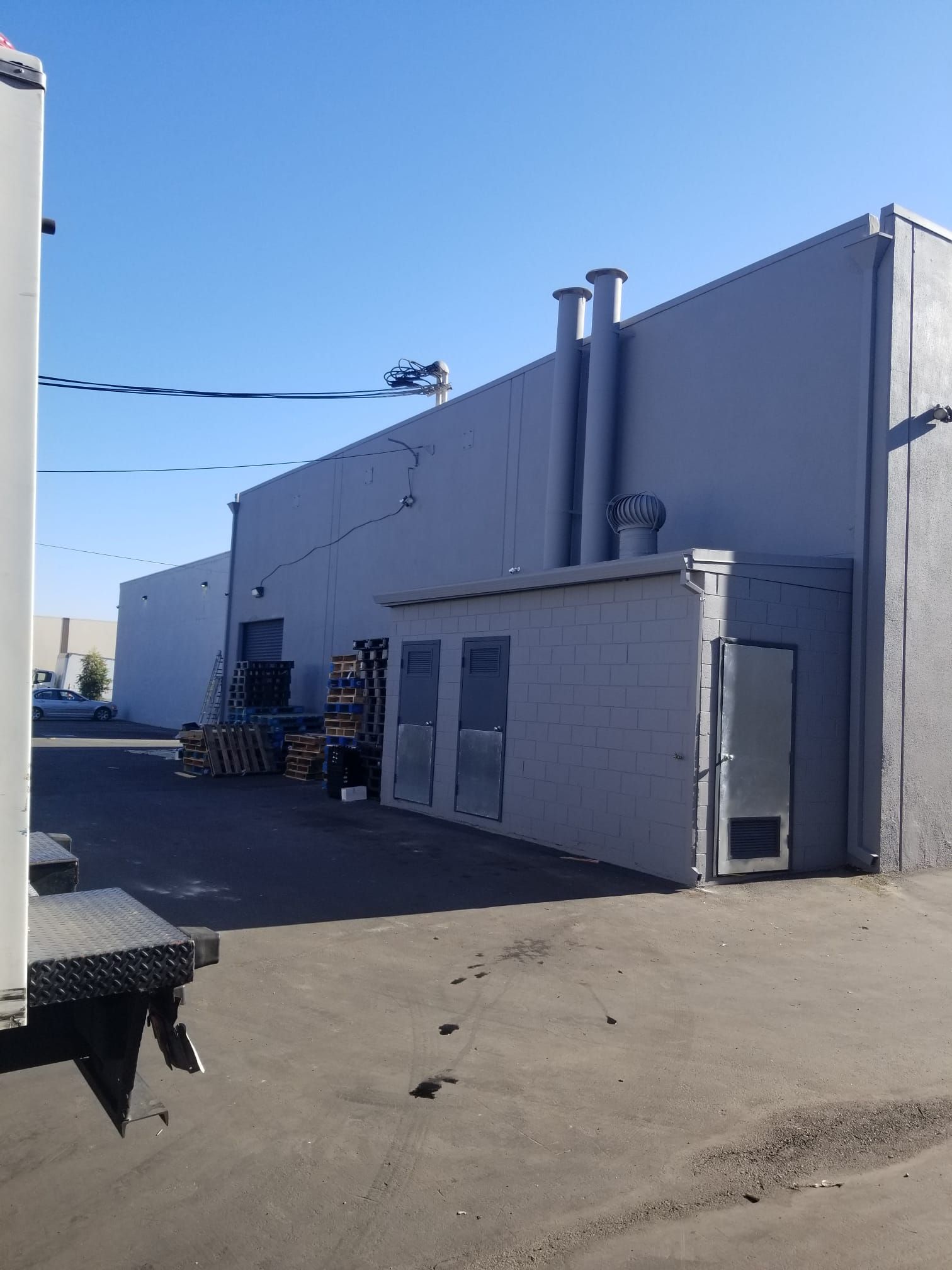 Industrial building exterior with gray walls, rooftop vents, and a paved loading area under a blue sky.