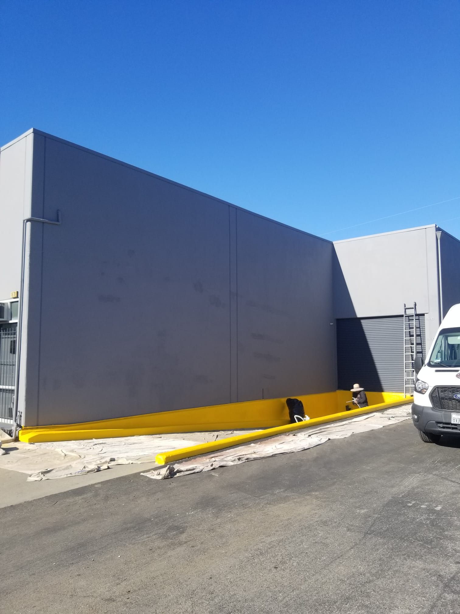 Gray industrial building with a yellow base beside a paved lot under a clear blue sky