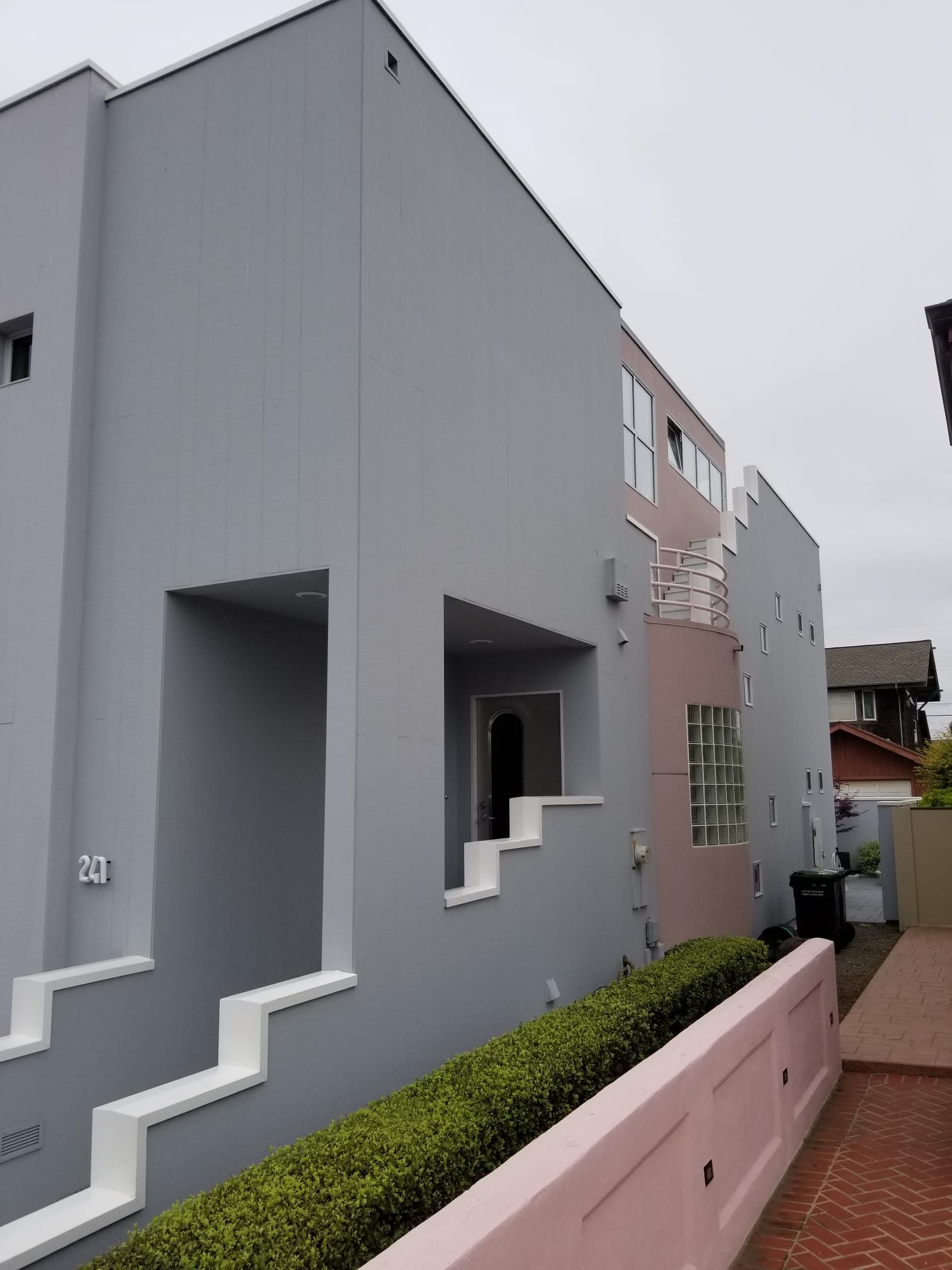 Gray building with geometric windows beside a pink wall and hedge on a brick path