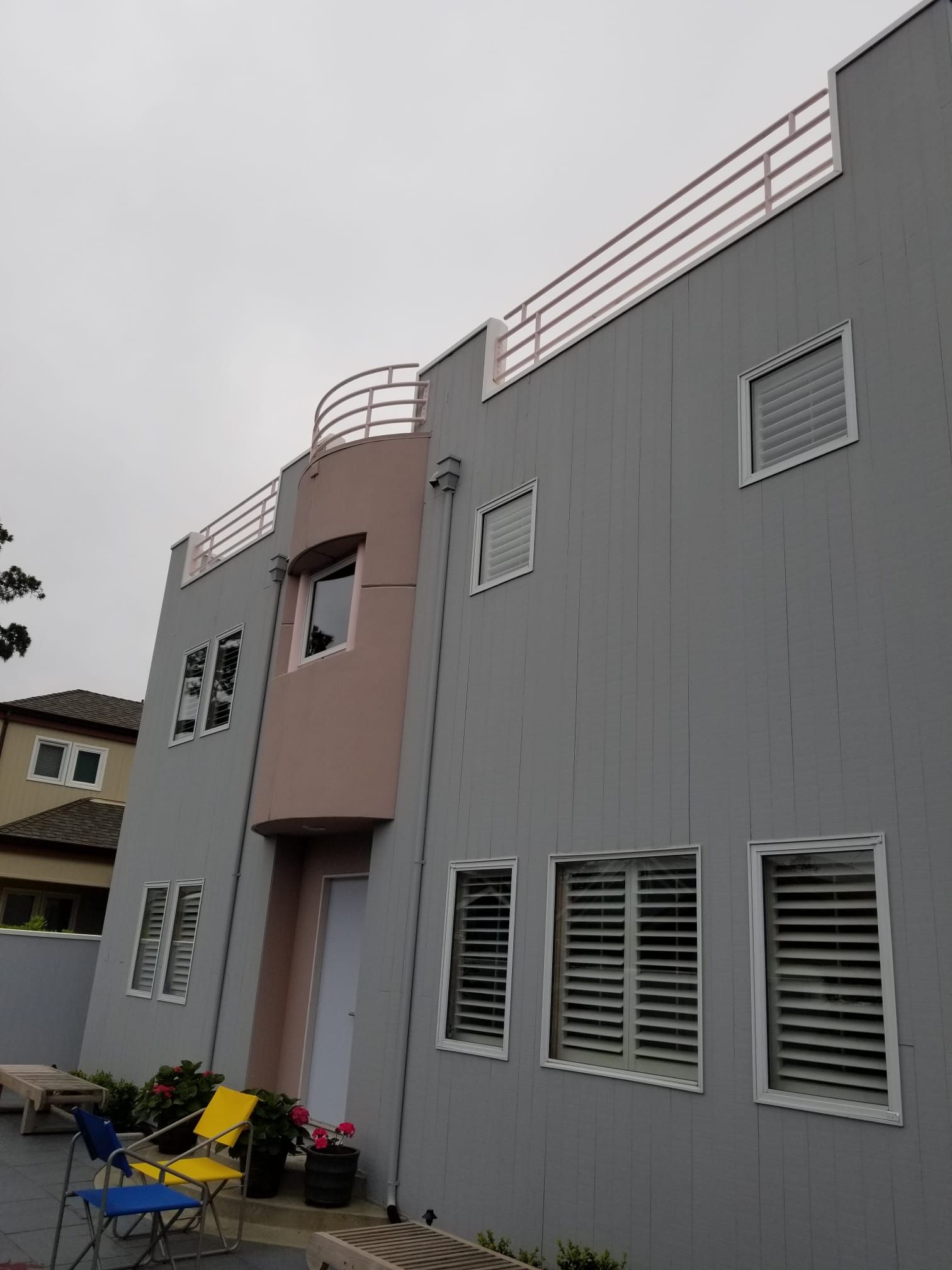 Modern gray and pink multi-story house with rooftop railings and louvered windows under an overcast sky