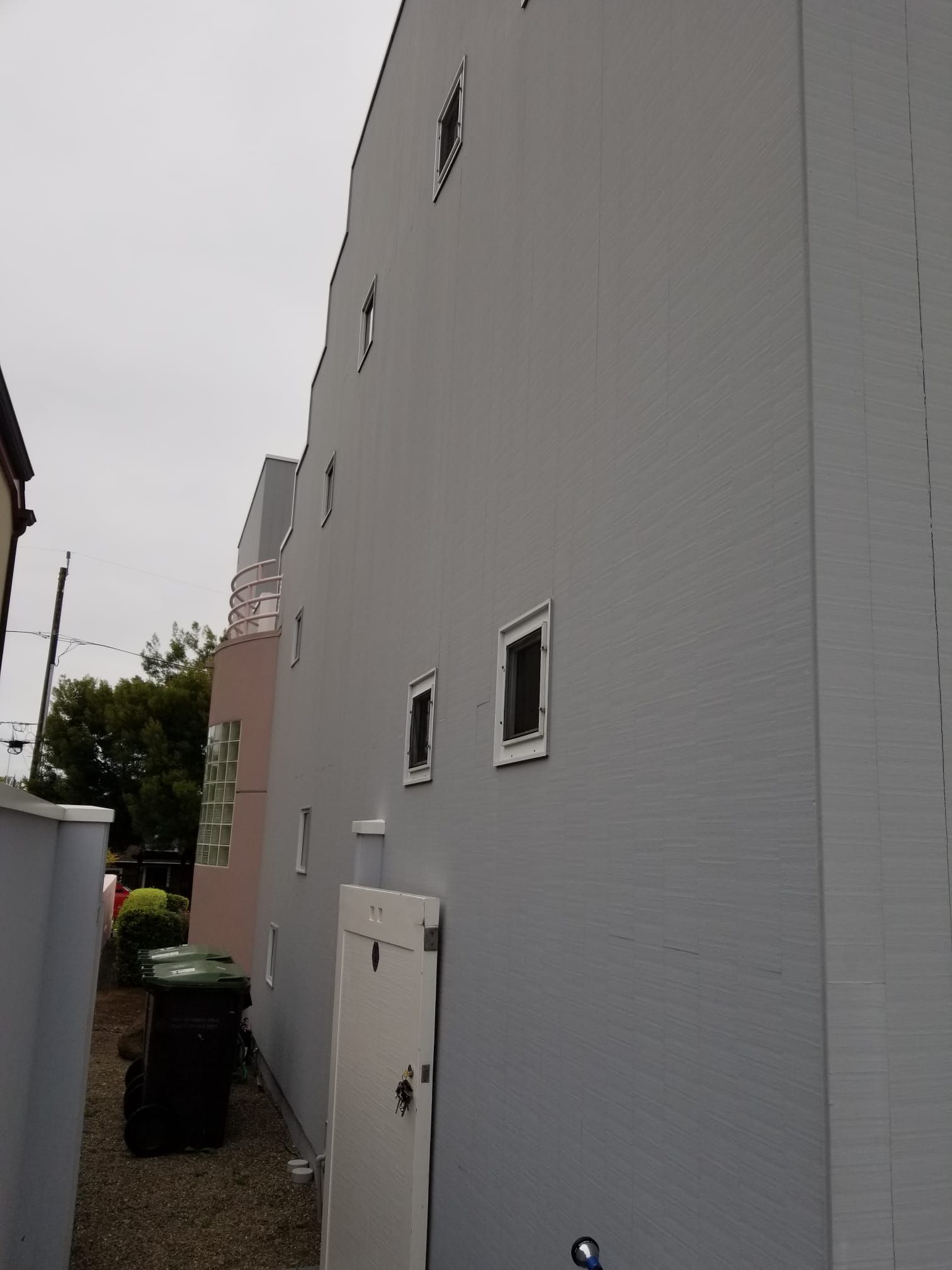 Narrow alley beside a gray building with small windows, a utility door, and trash bins.