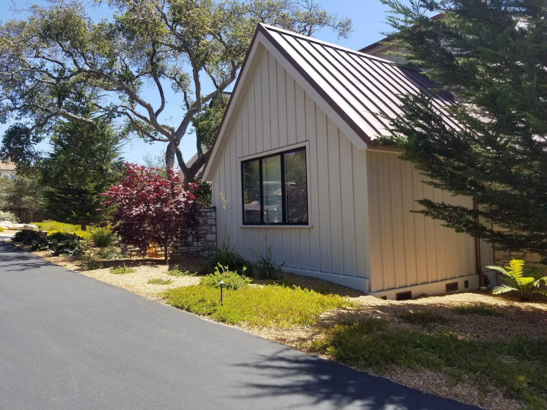 Small beige house with metal roof beside a paved road, surrounded by trees and shrubs.