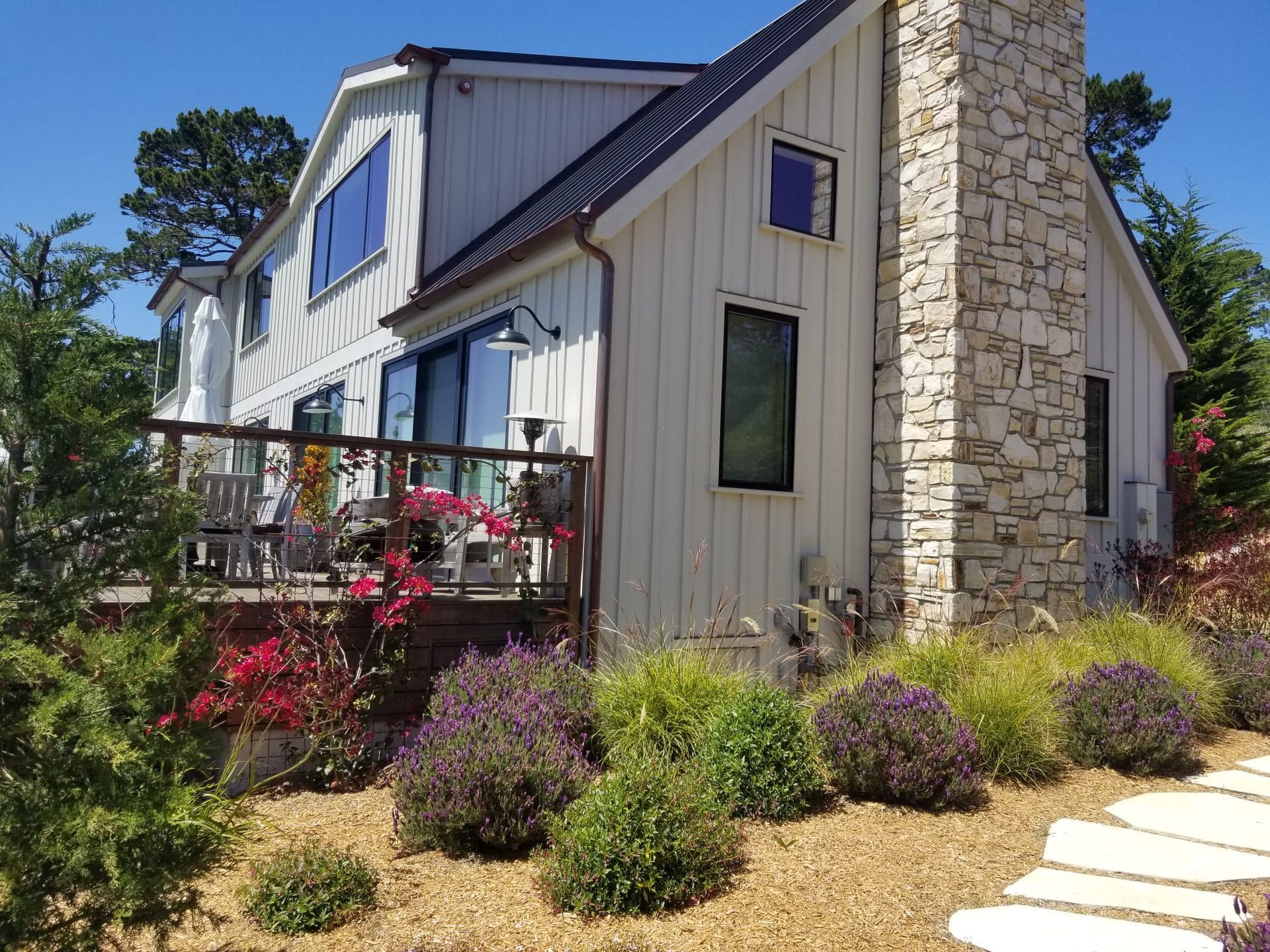 Modern house exterior with stone chimney, metal roof, and landscaped garden in bright sunlight