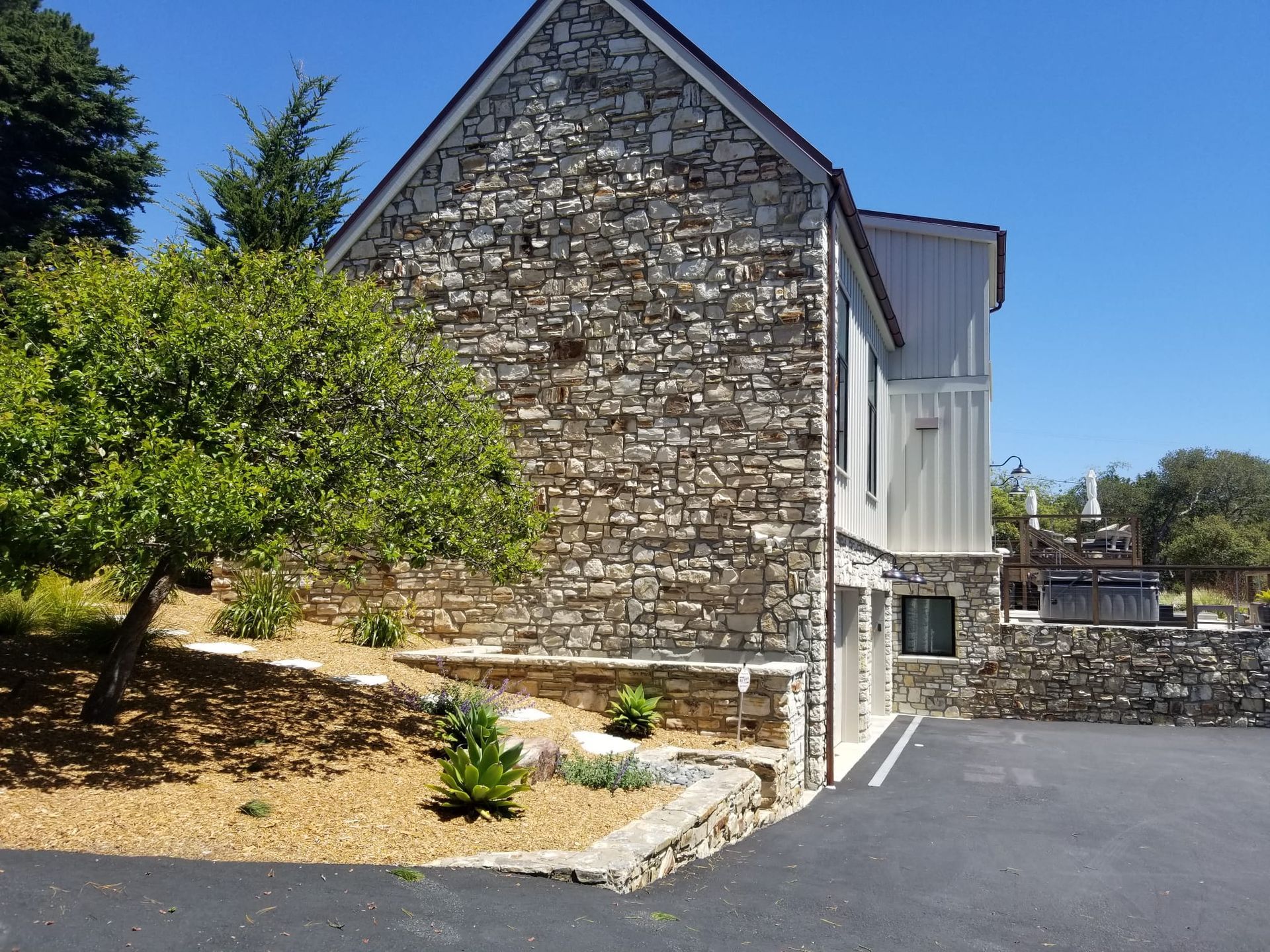 Stone-sided house beside a paved driveway, with a tree and dry landscaping under a clear blue sky
