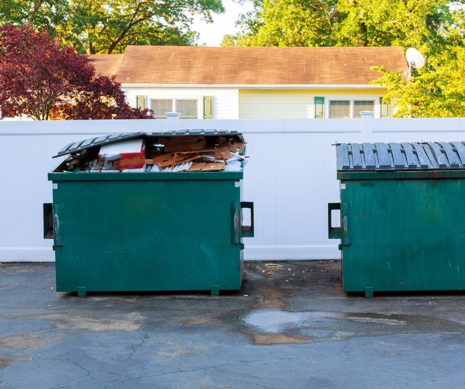 Two green dumpsters are in a parking lot in front of a house