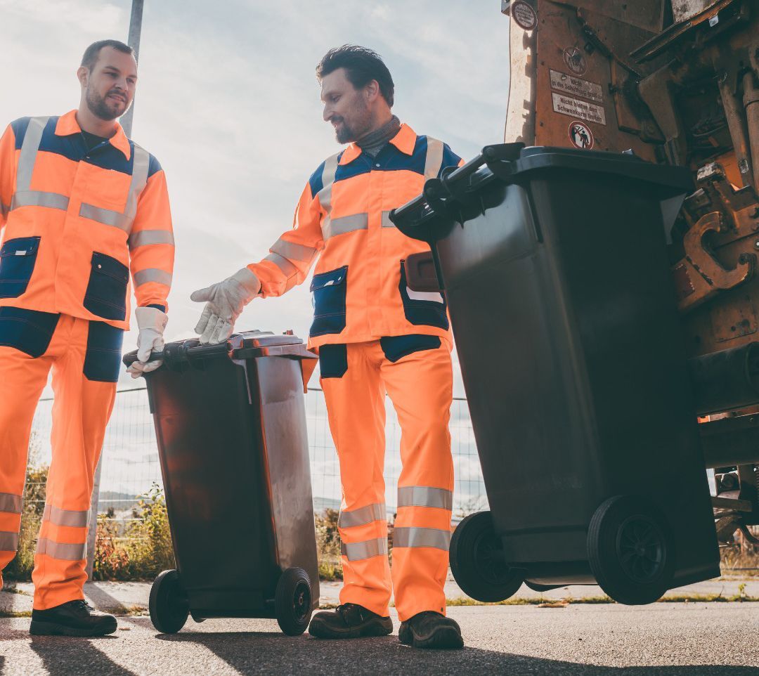 Two men are standing next to trash cans in front of a garbage truck.