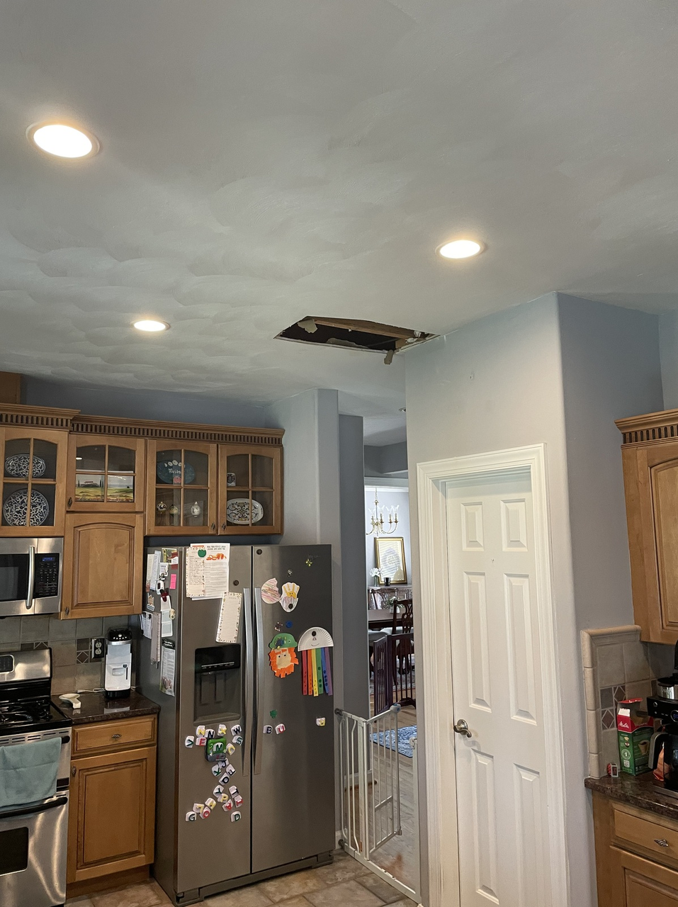 Kitchen with stainless steel fridge, wood cabinets, white door, and a damaged ceiling opening.