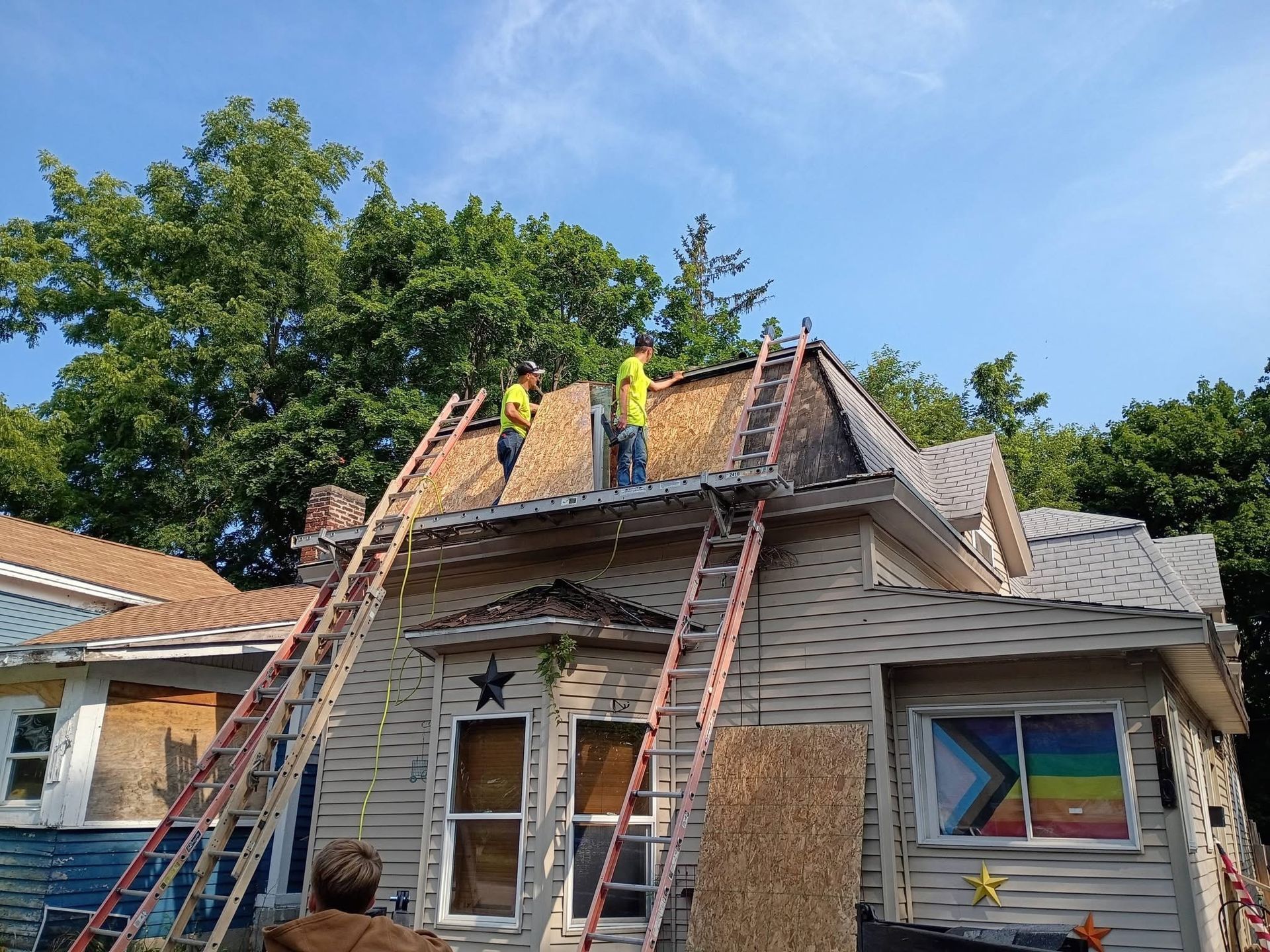 Roofers on a house roof with ladders, repairing shingles under a blue sky.