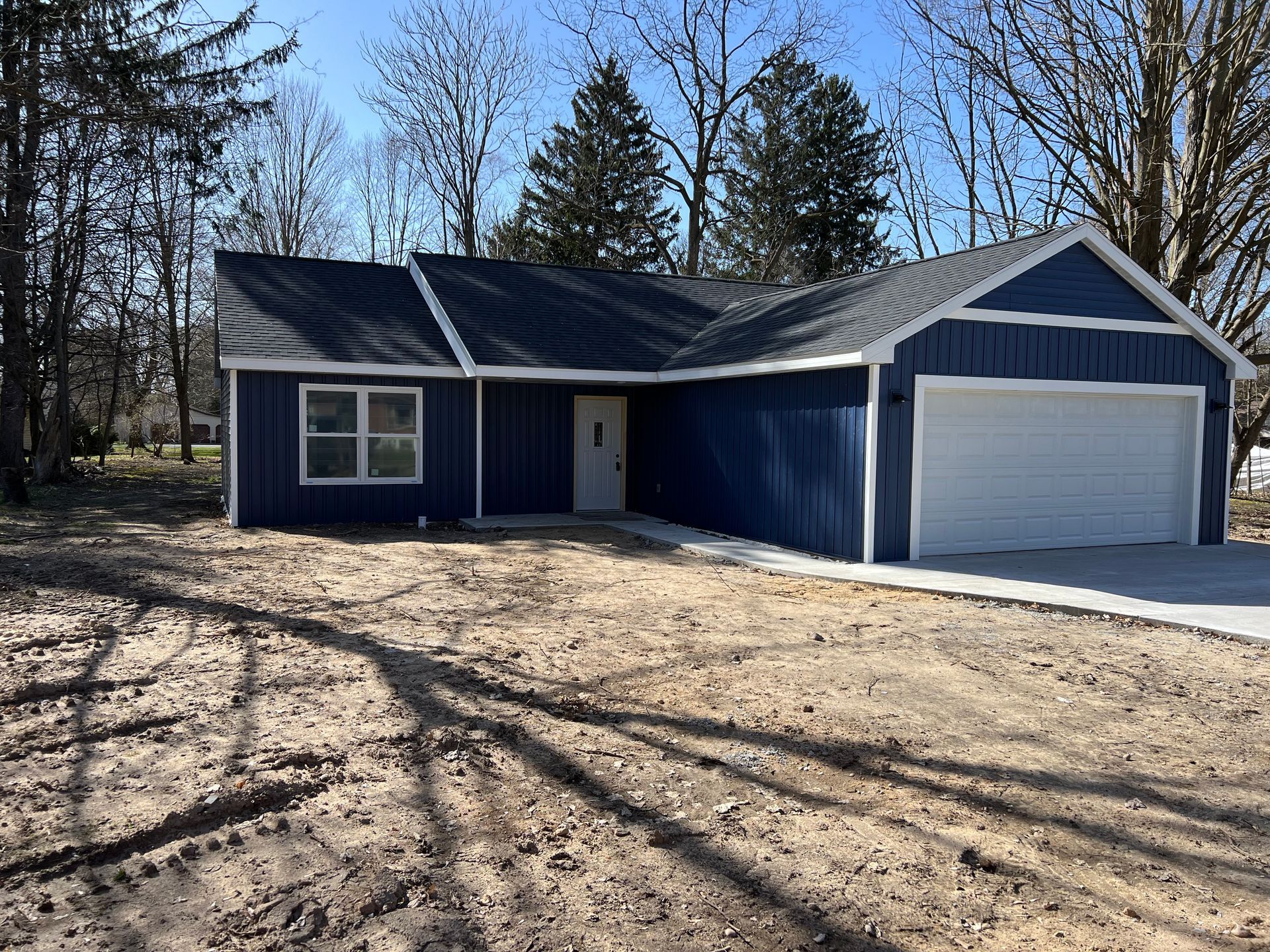 Blue house with white garage door and bare trees in a sandy yard on a sunny day