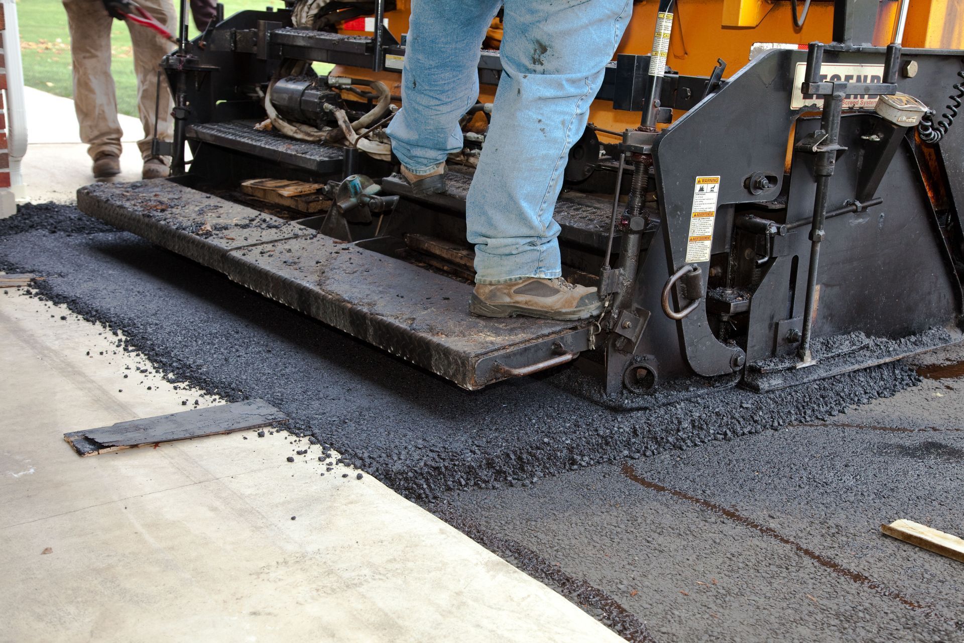 Man operating asphalt paving machine, laying down fresh asphalt on a concrete surface.