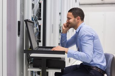 A man is sitting at a desk in front of a laptop computer in a server room.