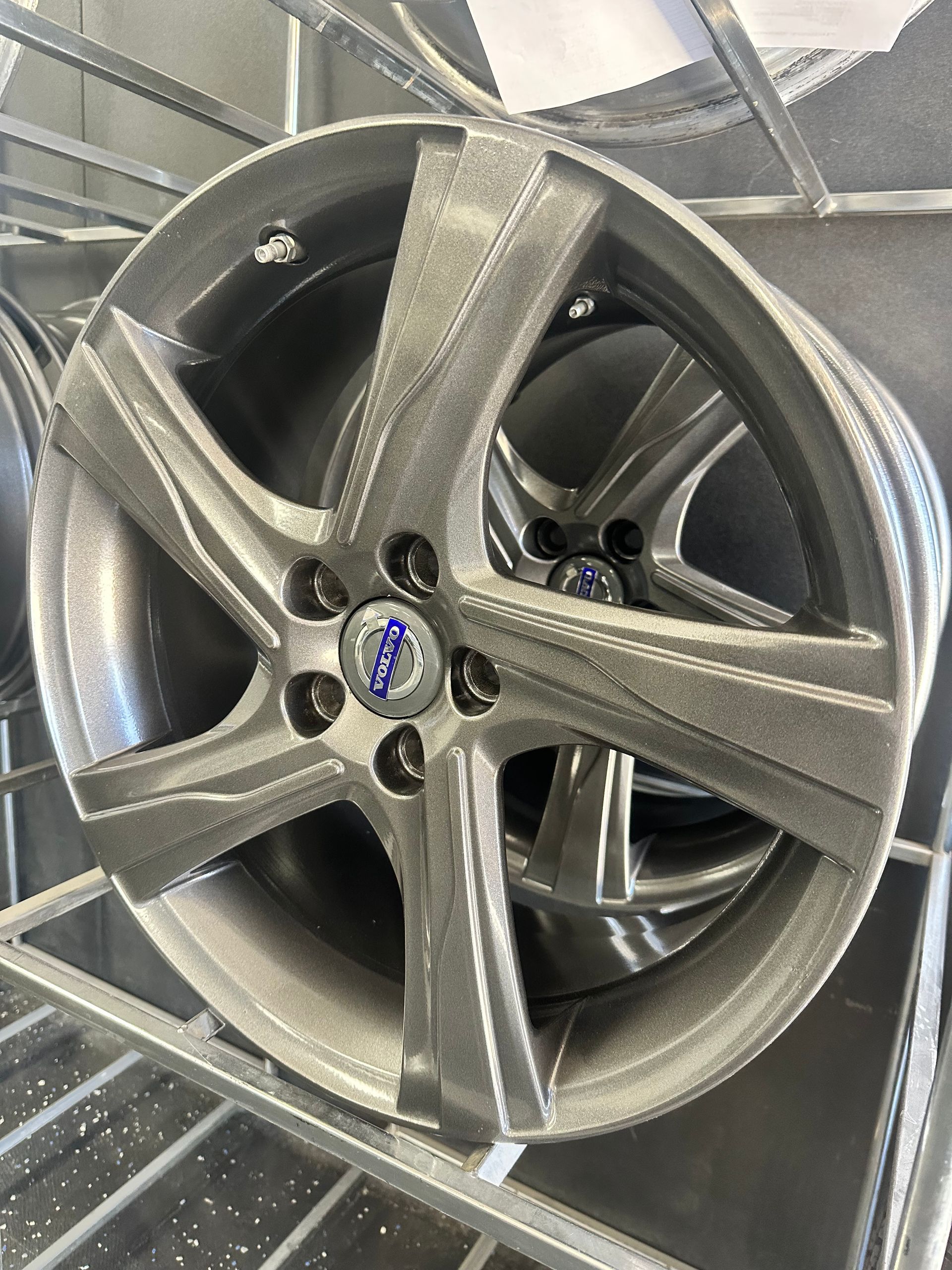 Gray Volvo car wheel with a five-spoke design, resting on a metal rack. The center cap displays the Volvo logo.