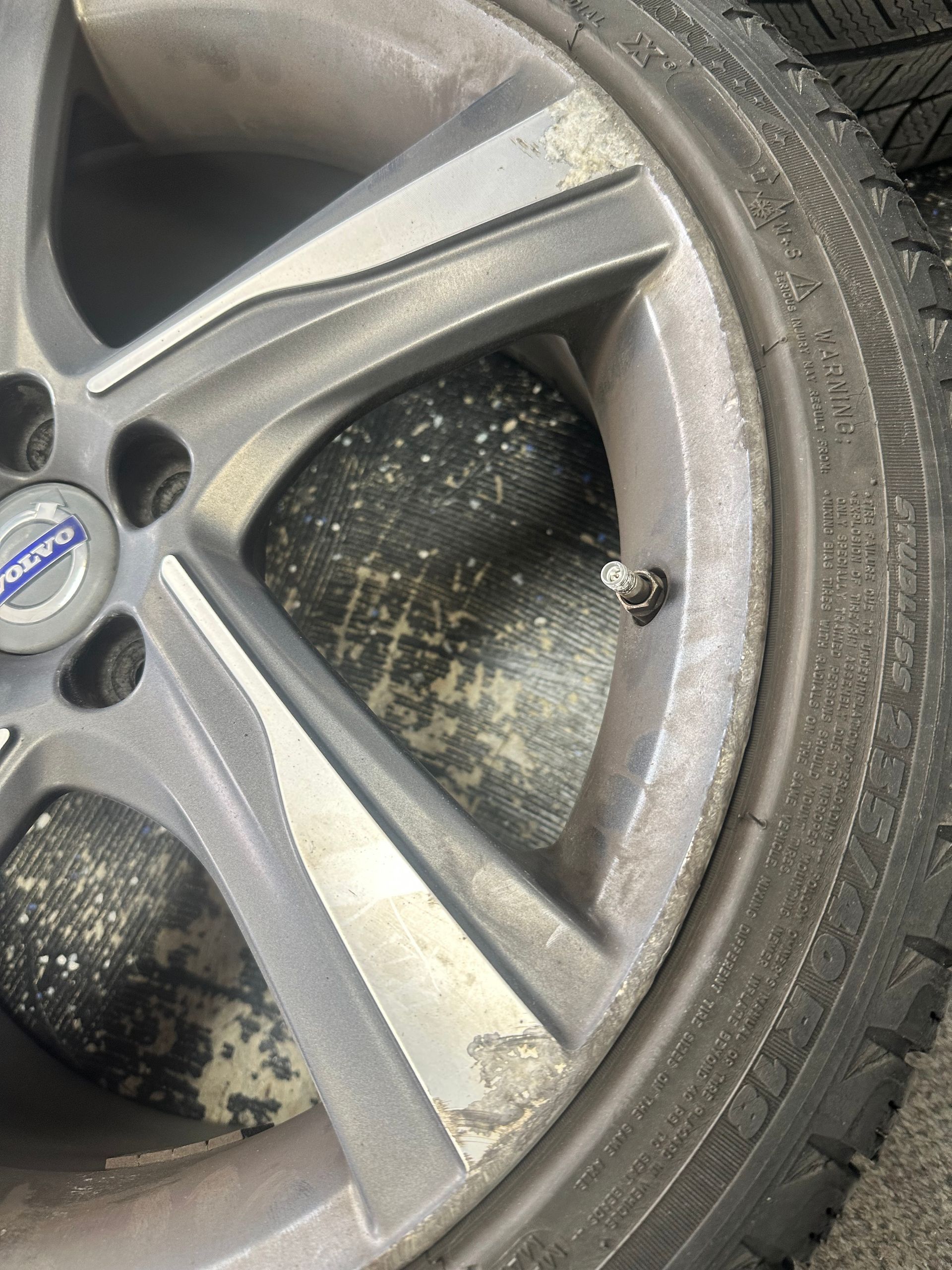 Close-up of a dirty Volvo car wheel. The rim is gray, with the Volvo logo at the center. The tire is black and has snow.