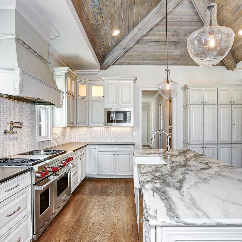 Elegant white kitchen with marble island, wood ceiling, and stainless steel appliances.