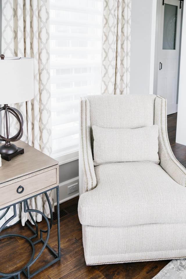 Beige chaise lounge next to a side table with a lamp, in a bright room with patterned curtains.