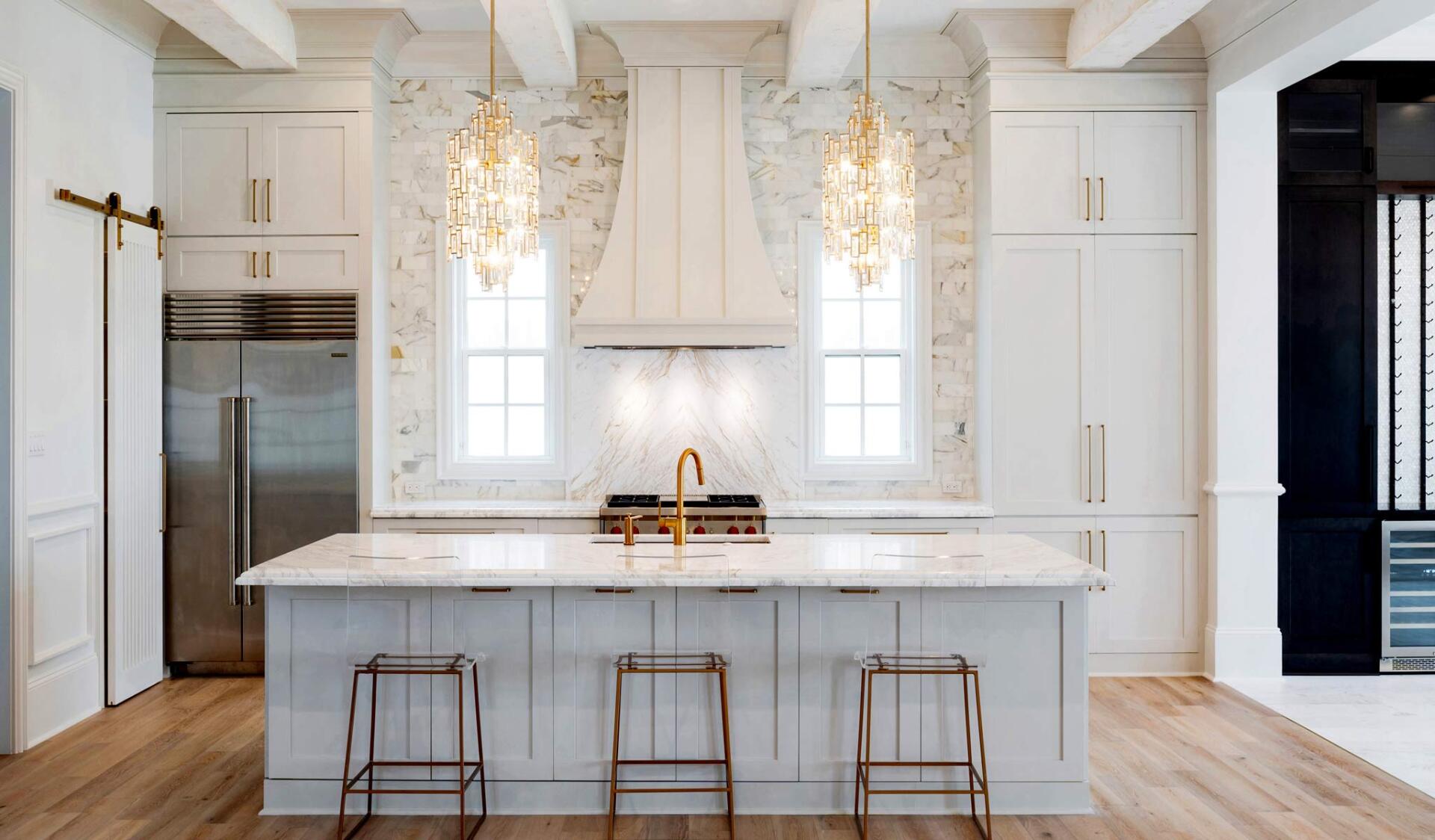 Elegant white kitchen with marble island, gold accents, and two chandeliers.