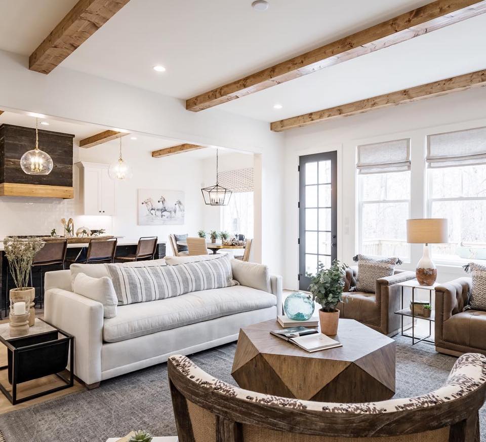 Spacious living room with white walls, wooden beams, and neutral furniture. Kitchen visible in the background.