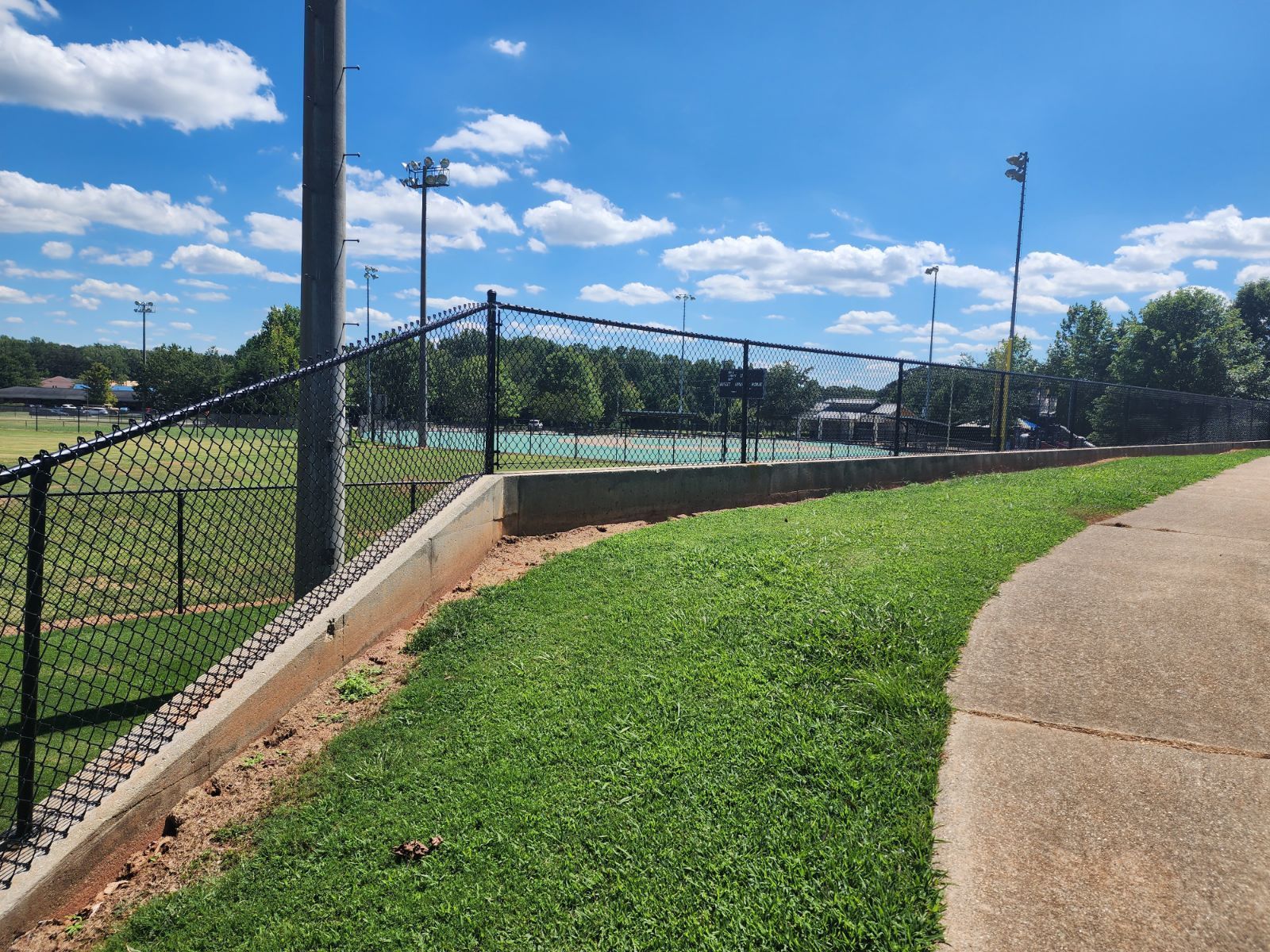 A chain link fence surrounds a tennis court in a park.