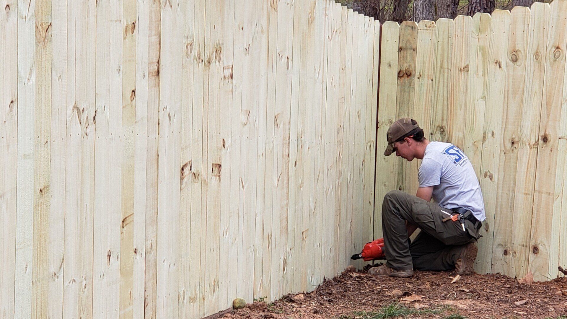 A man is kneeling down next to a wooden fence.