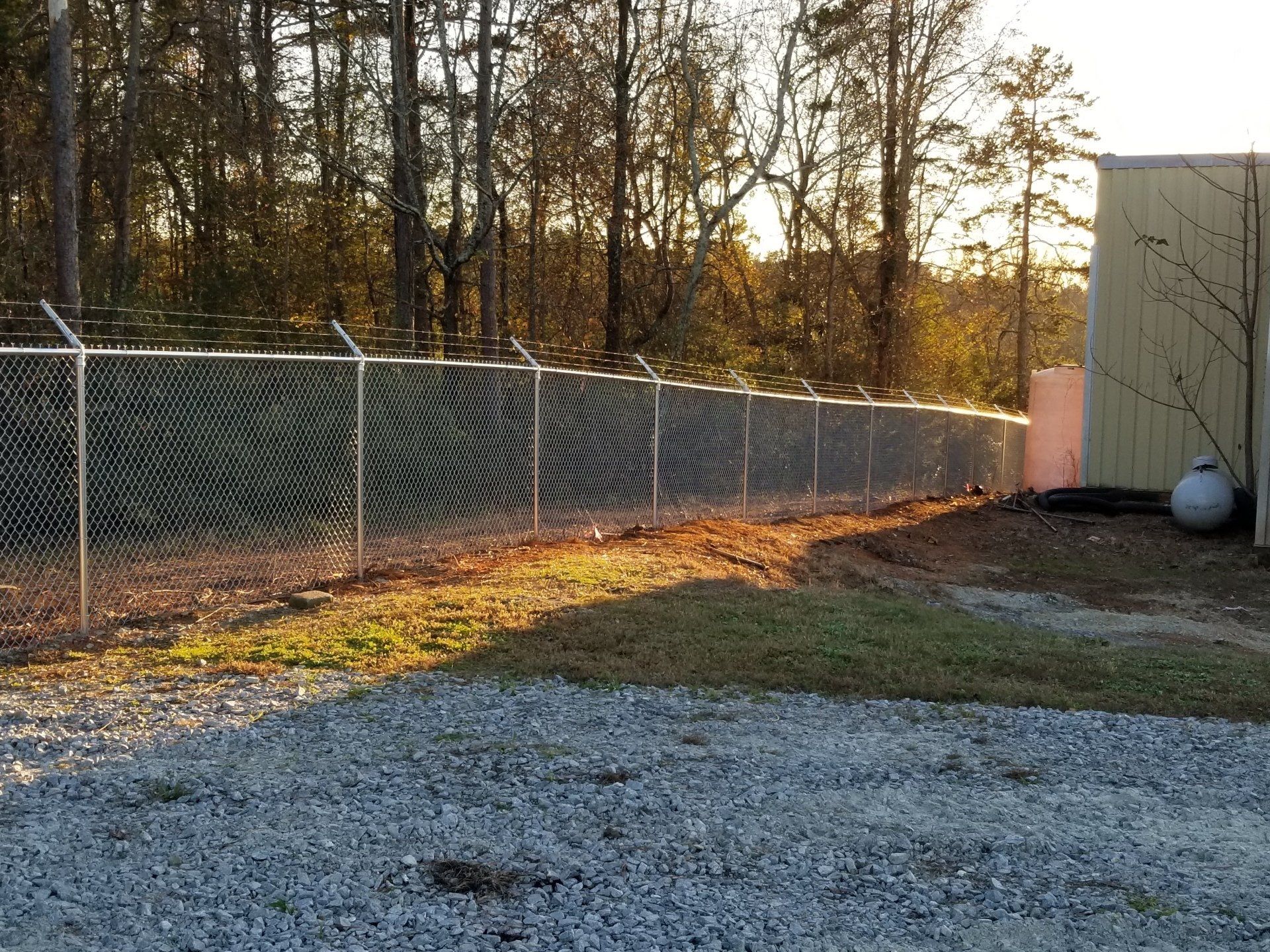 A chain link fence is surrounding a gravel area with trees in the background.