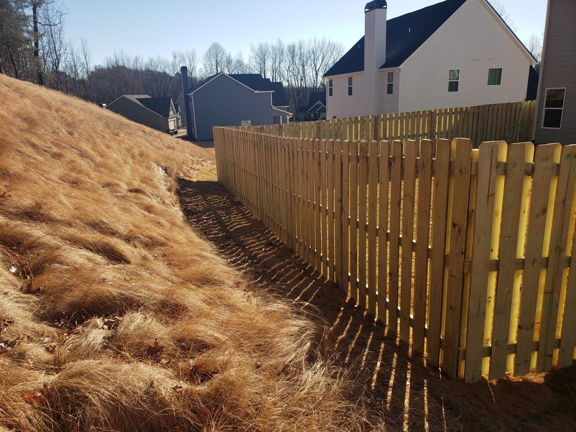 A wooden picket fence surrounds a dry grassy hillside in front of a house.