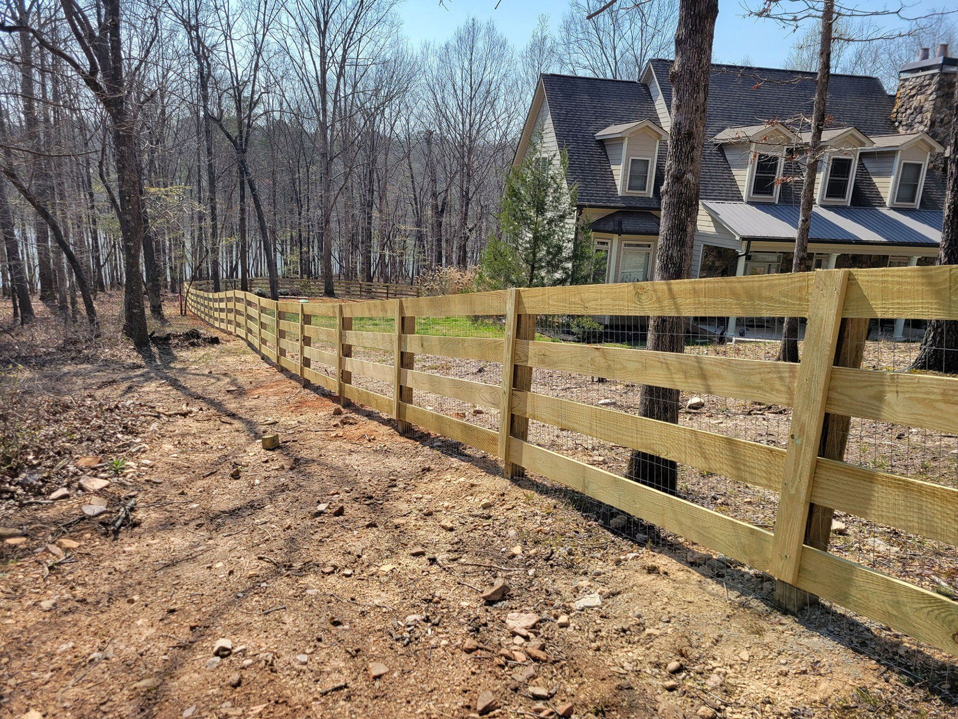 A wooden fence surrounds a dirt path leading to a house in the woods.