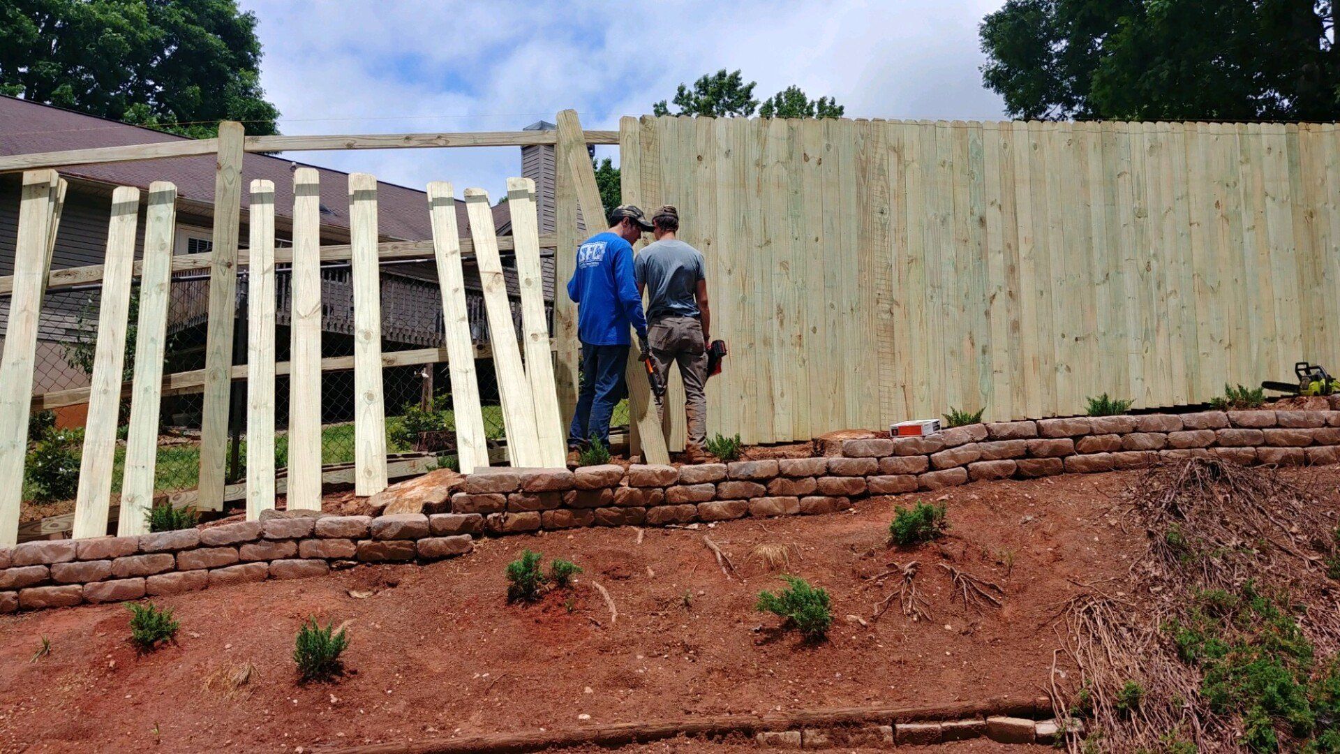 Two men are standing in front of a wooden fence