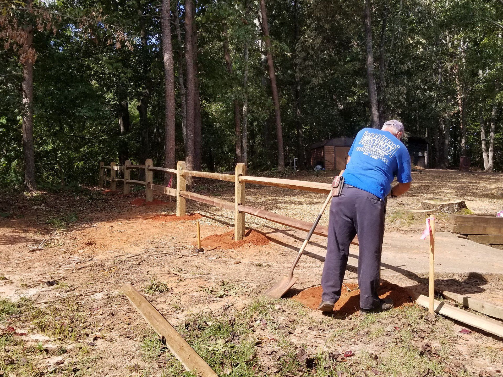 A man in a blue shirt is raking dirt in front of a wooden fence.