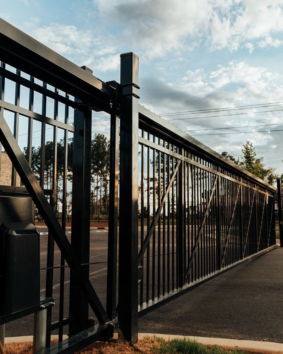 A black metal fence surrounds a parking lot with trees in the background