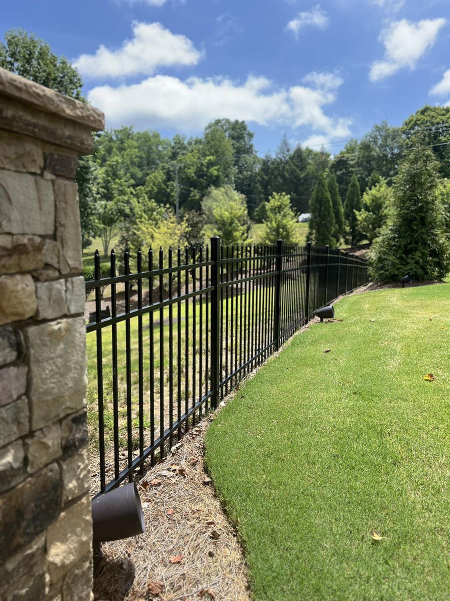 A black metal fence surrounds a lush green field next to a stone wall.