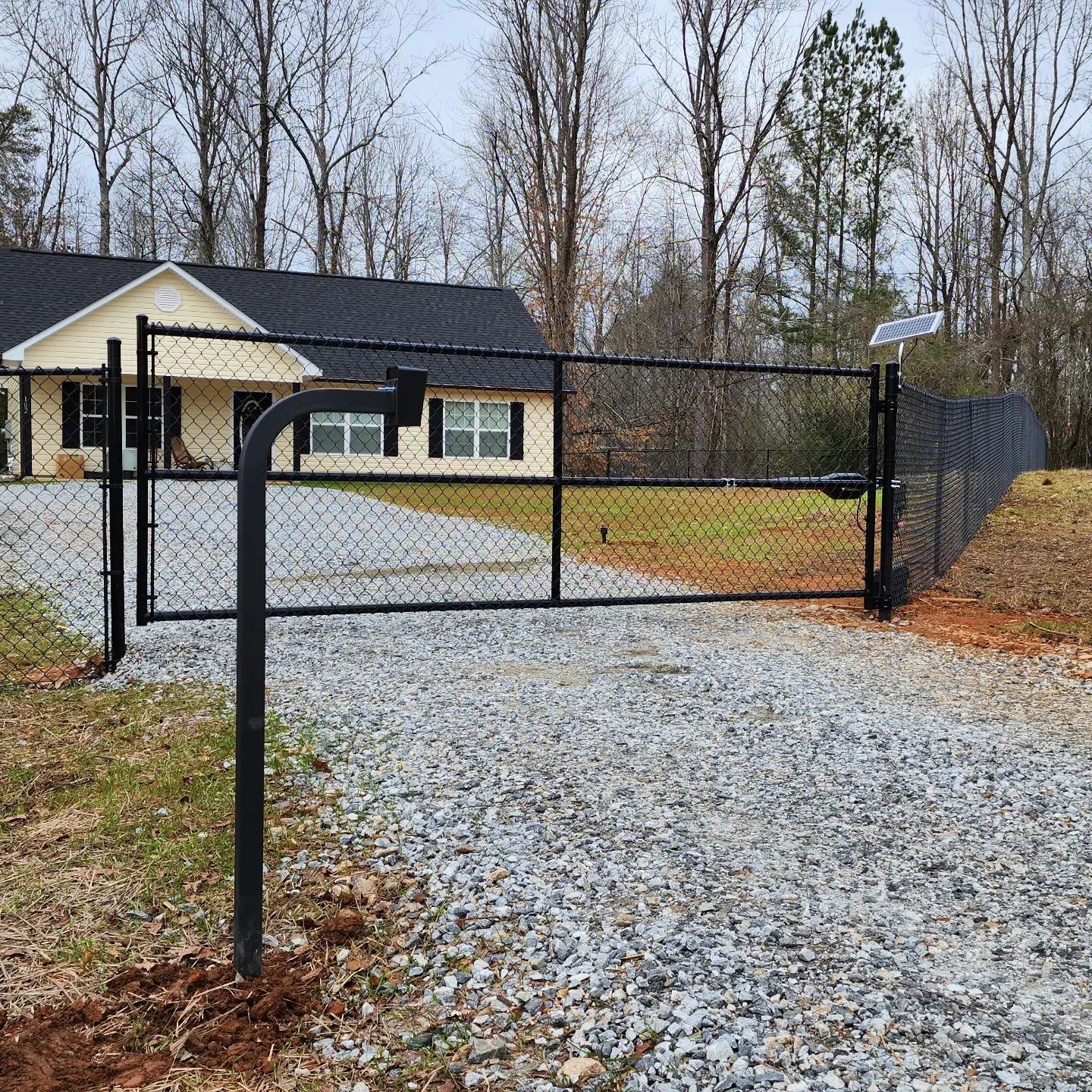 A chain link fence surrounds a gravel driveway leading to a house.