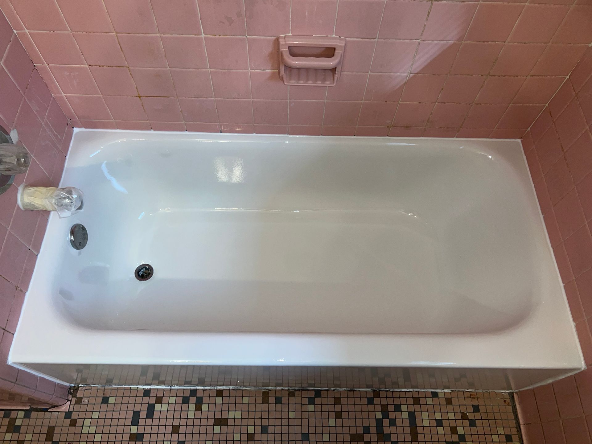 White bathtub in a pink-tiled bathroom, with a soap dish above and a patterned floor.