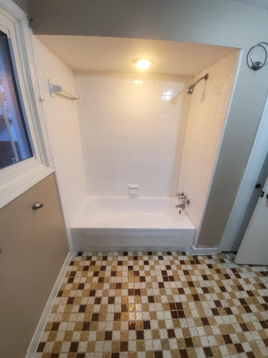 White bathtub and shower unit in a bathroom with brown and beige tile floor and a window.