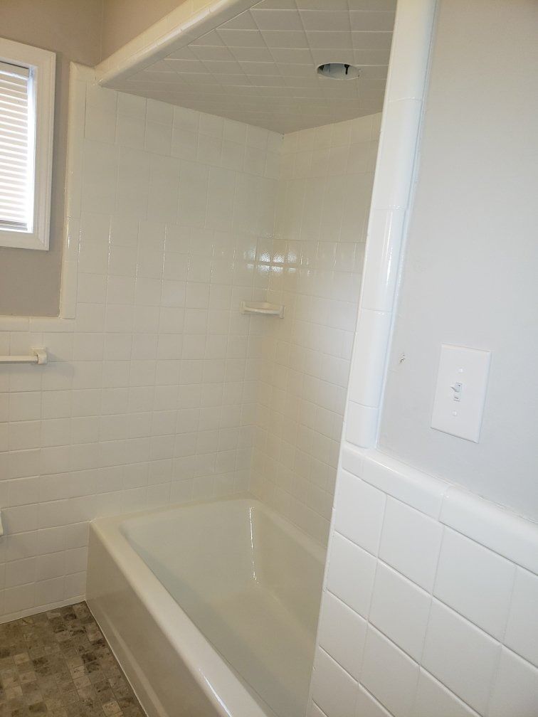 White bathroom with a tub and shower, tiles on the walls, and a window with blinds.