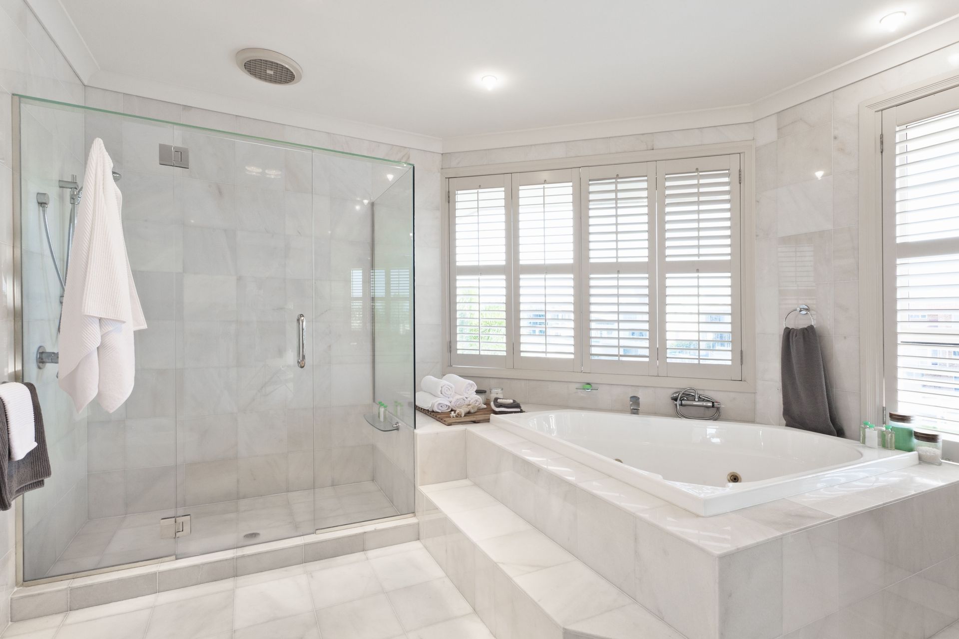 Spacious white bathroom with glass shower, soaking tub, and window with shutters.