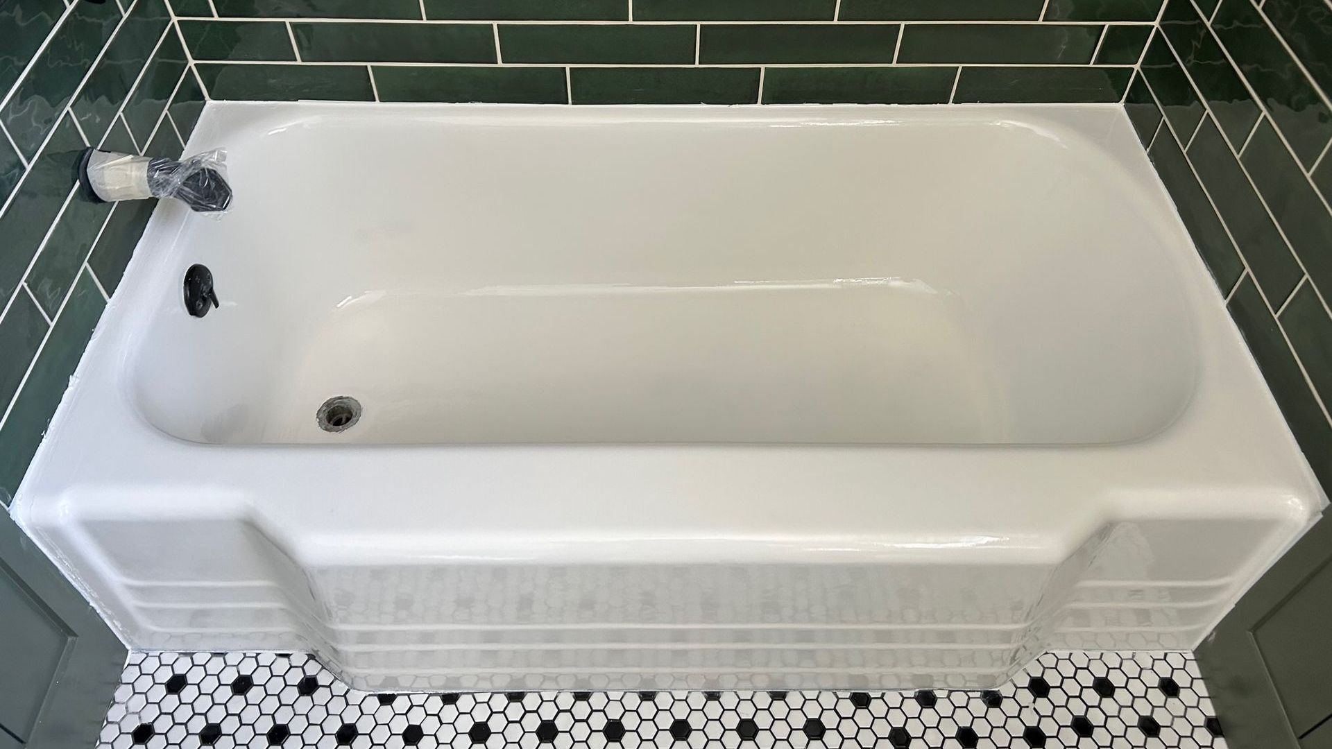White bathtub with black fixtures, surrounded by green tile and black and white mosaic floor.