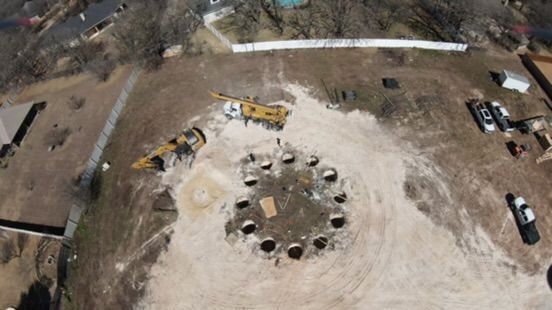 An aerial view of a construction site featuring a circular pattern of drilled holes in the ground with heavy machinery.